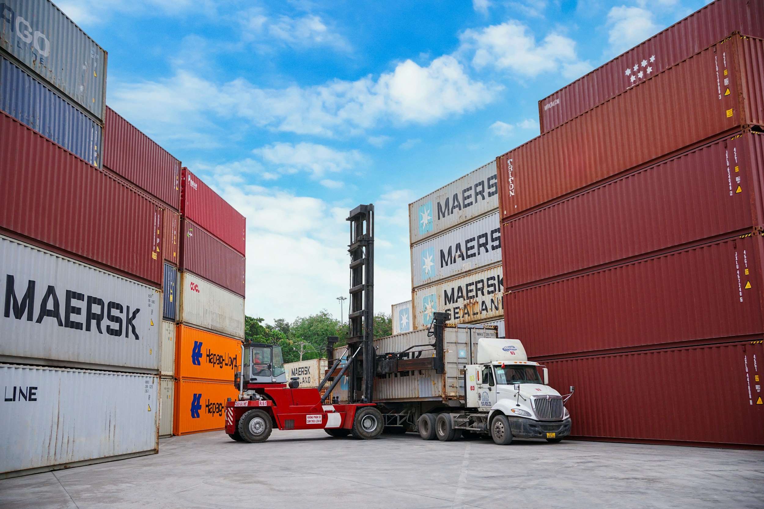 A large shipment of stacked shipping containers in a port yard. Several containers display the MAERSK logo, and a forklift is lifting one container while a semi-truck is parked beneath. The sky is partly cloudy.