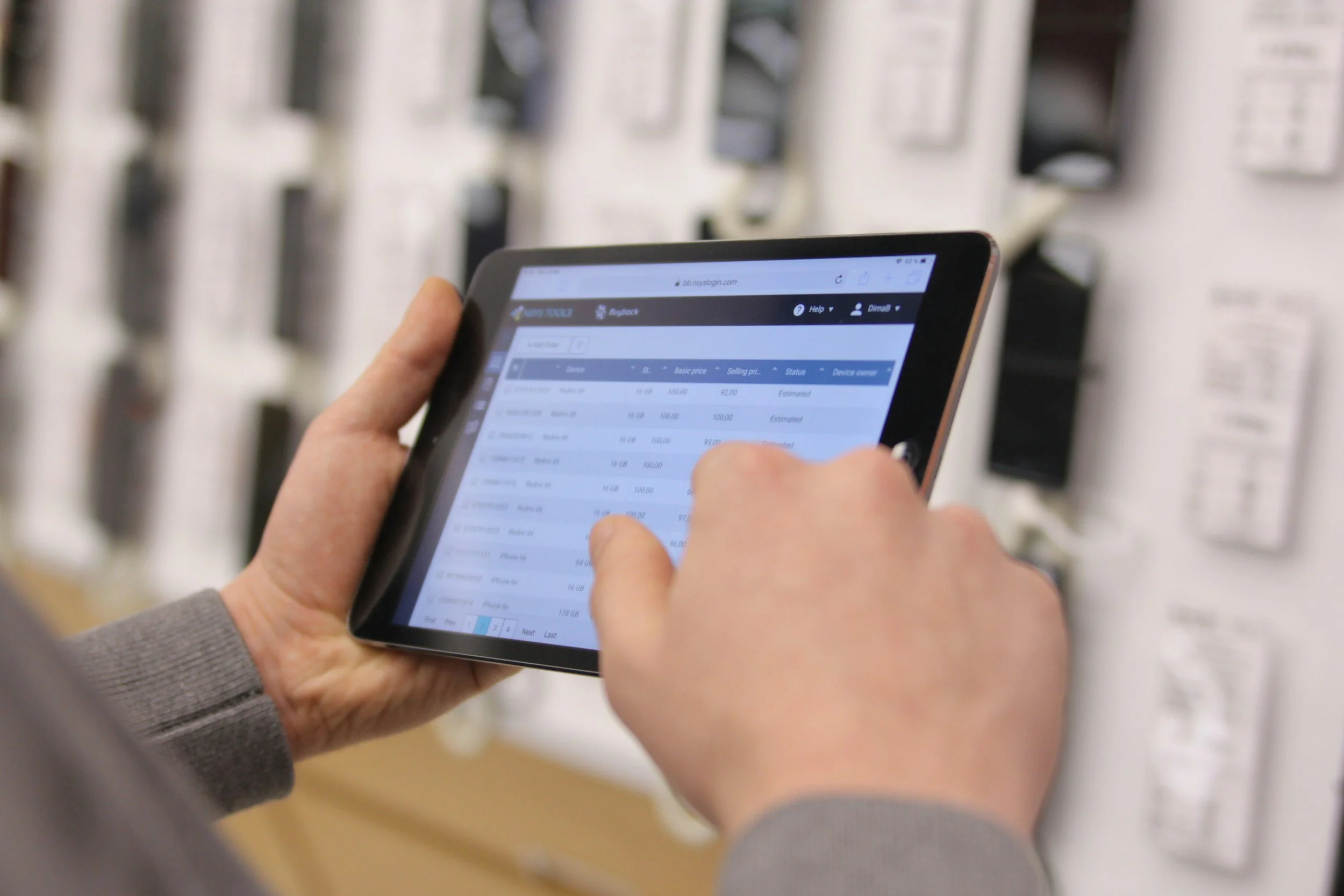 Person holding a tablet device displaying a spreadsheet, with shelves of electronic devices or accessories in the background.