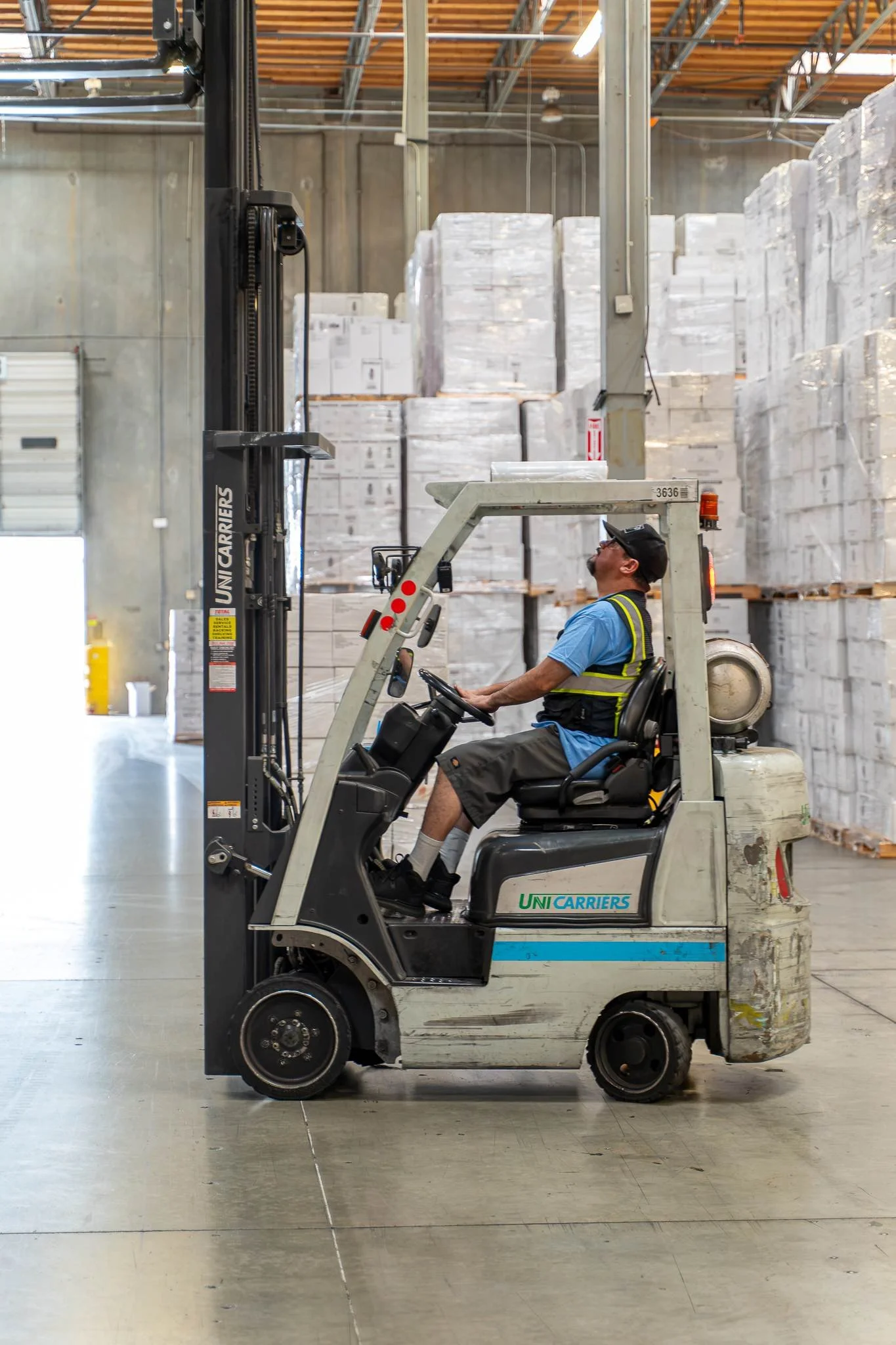 A man wearing a blue shirt, safety vest, and cap driving a warehouse forklift amid stacked pallets of goods in a warehouse.