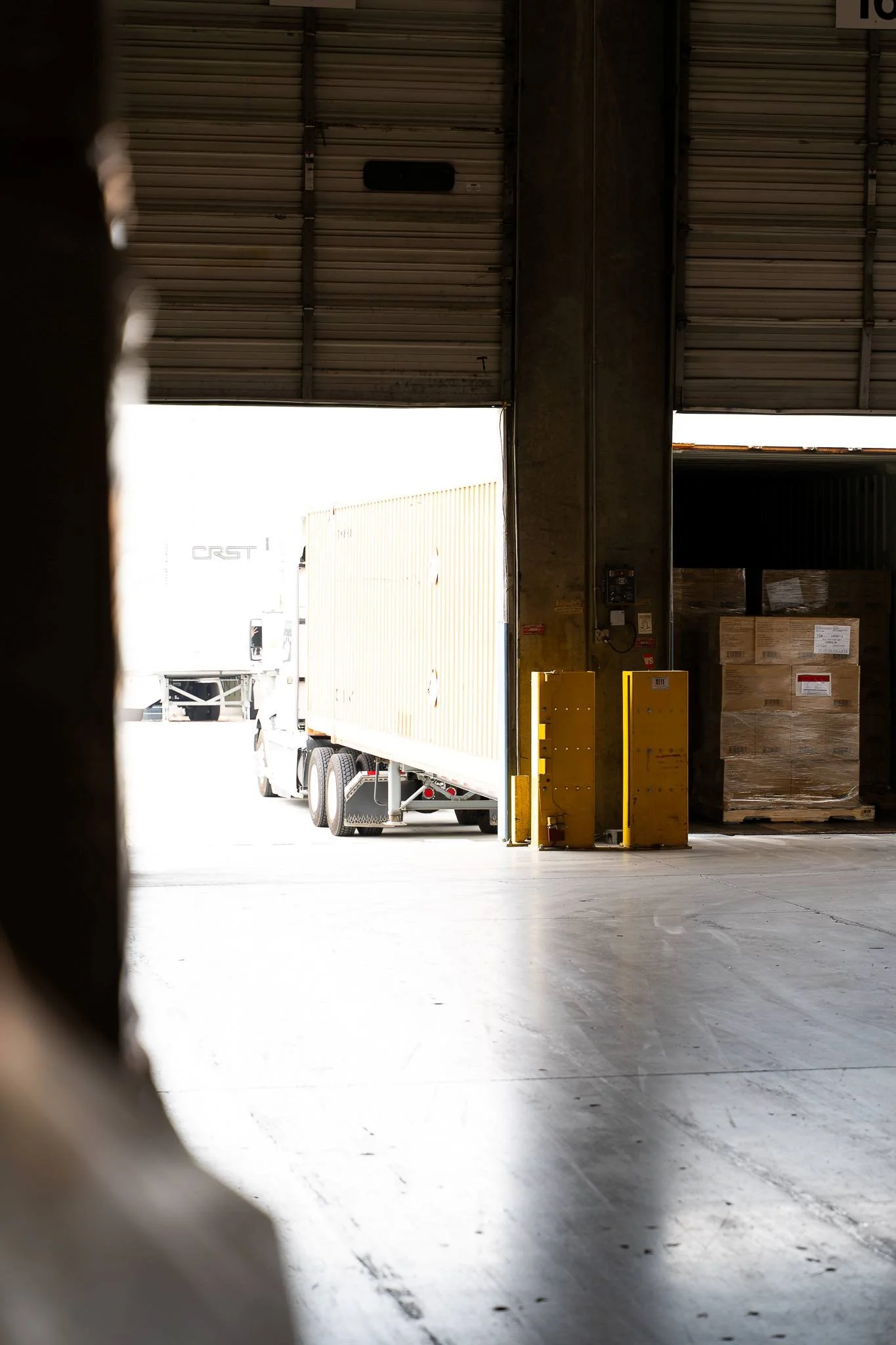 View of a loading dock with a semi-truck and stacked boxes inside a warehouse, seen through an open bay door.