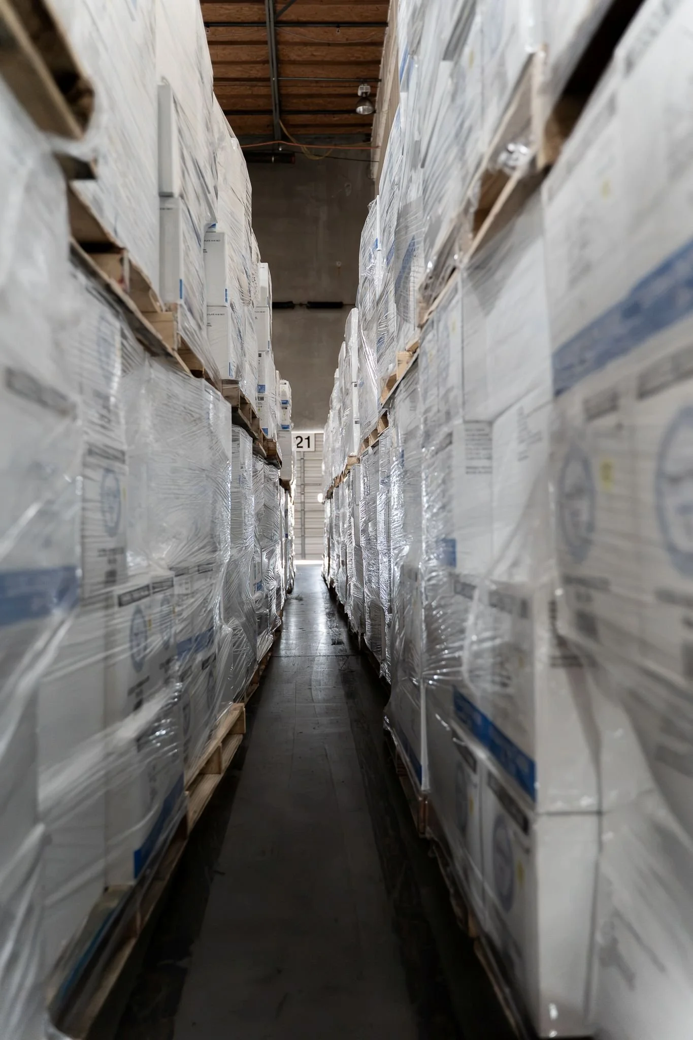 A narrow aisle in a warehouse with tall shelves on each side, stacked with boxes wrapped in plastic.