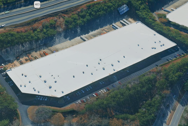 Aerial view of a large rectangular retail or warehouse building with a white roof, surrounded by parking lots and trees, with a highway nearby.