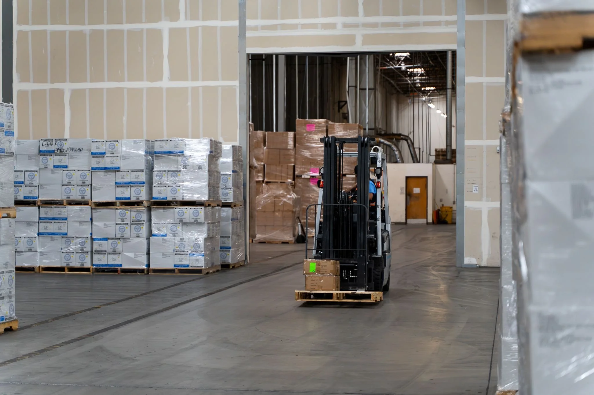 An indoor warehouse with stacks of boxed products on pallets, a forklift, and unfinished walls.