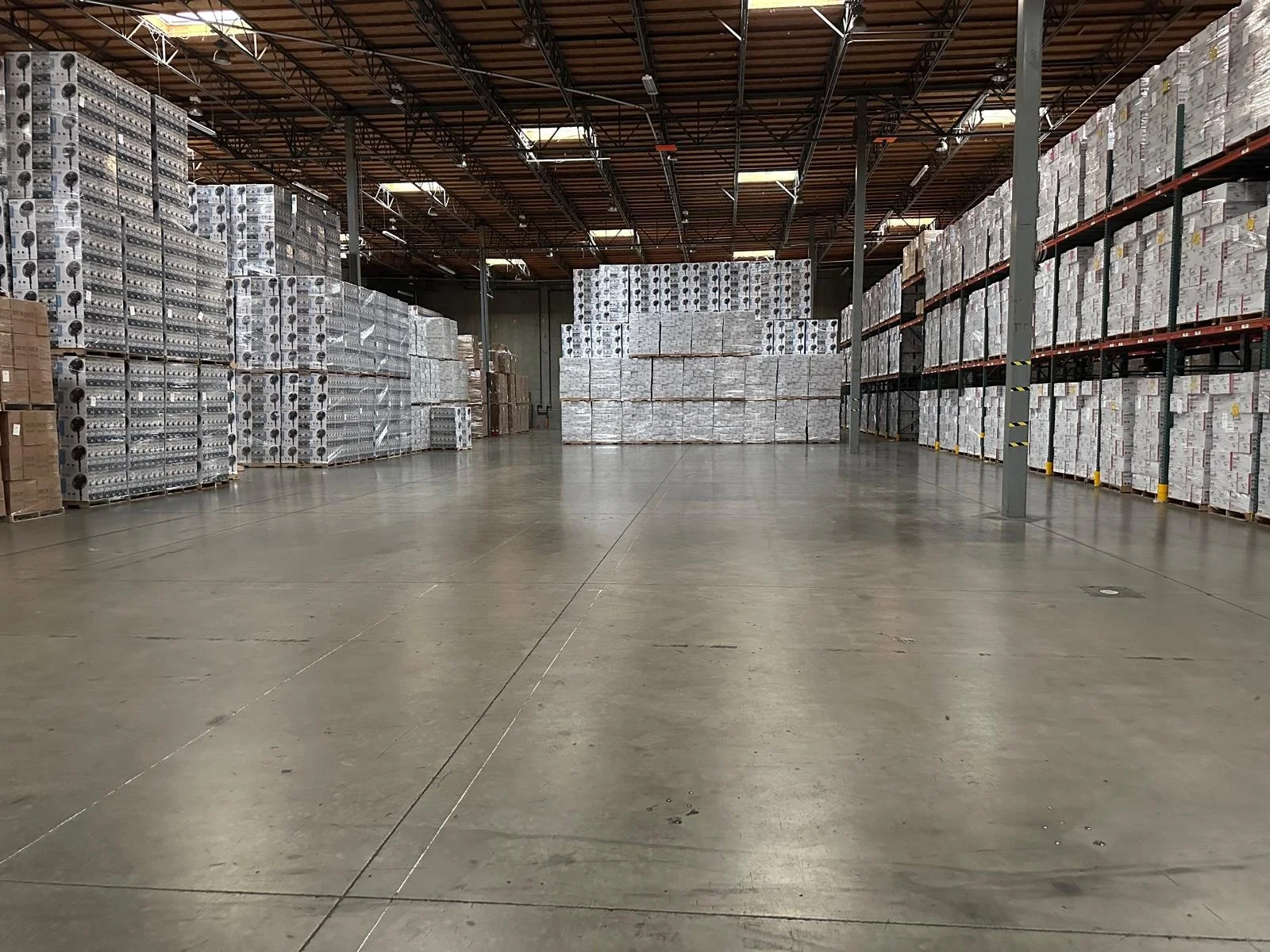 Interior of a warehouse with stacked pallets of boxed goods on the right and left sides, and a large open space in the center.