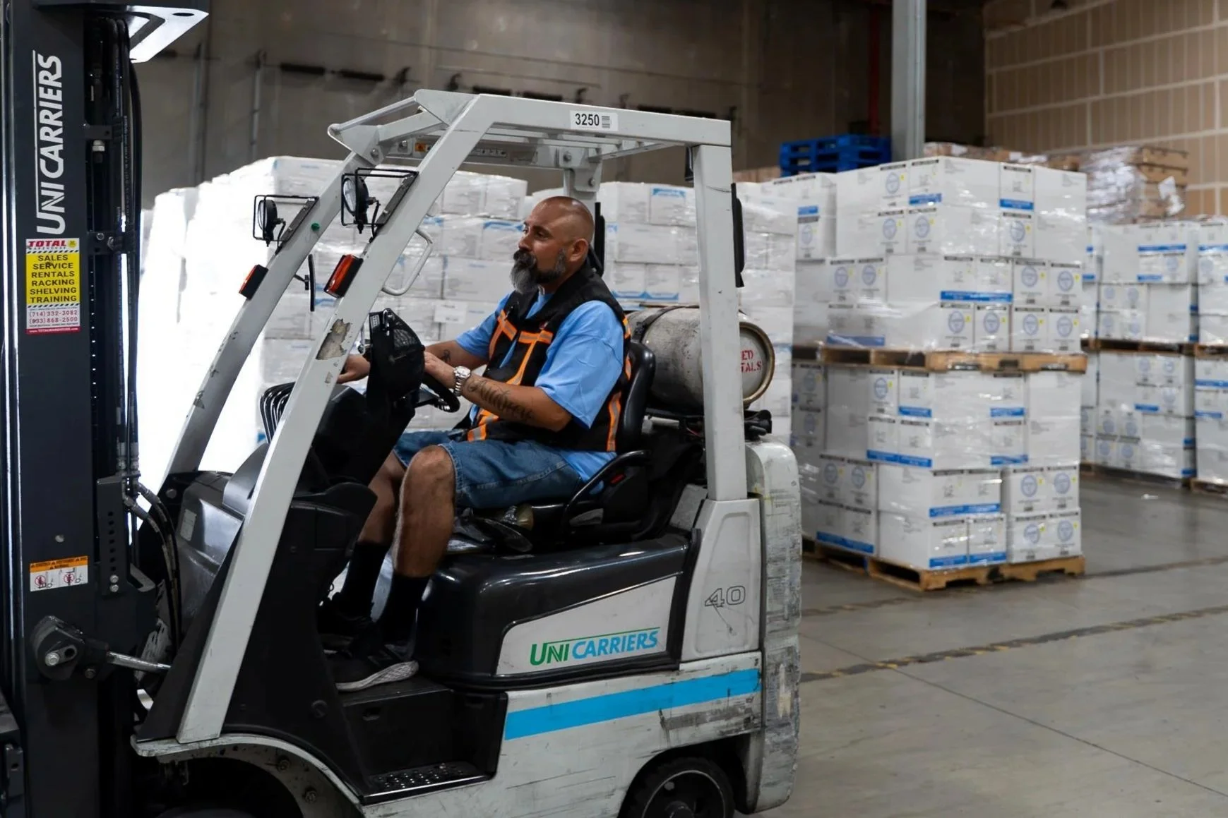A man operating a forklift inside a warehouse, surrounded by stacks of boxed goods on pallets.