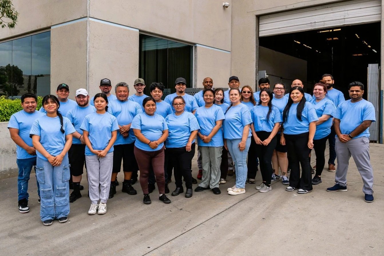 Group of people wearing matching blue T-shirts standing outdoors in front of a building, smiling at the camera.