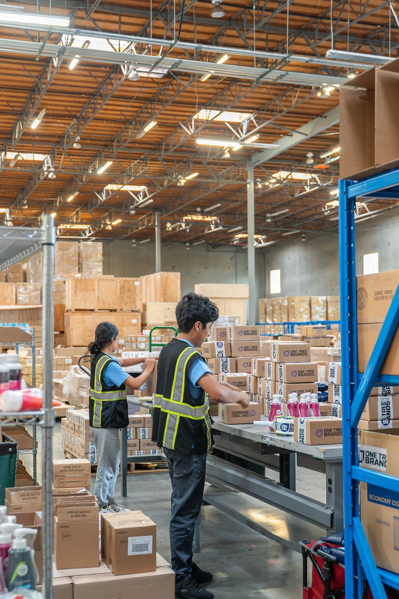 Workers wearing reflective vests packing boxes of products in a warehouse with high ceilings, wooden beams, and large windows.