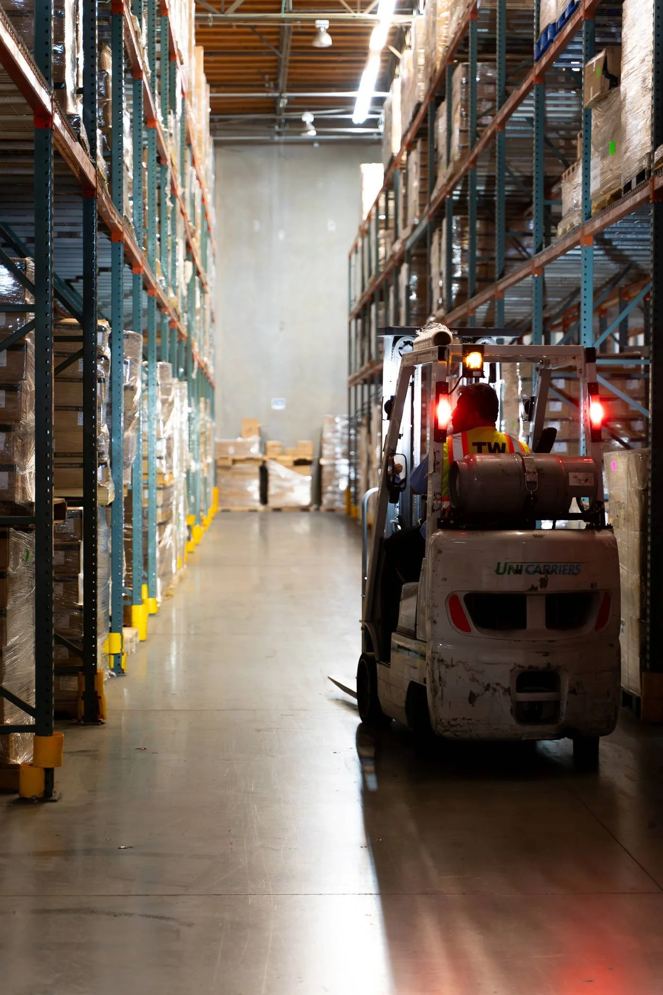 A warehouse aisle filled with shelves stocked with brown boxes. A person operating a small forklift is moving through the aisle, with sunlight streaming in from the ceiling.