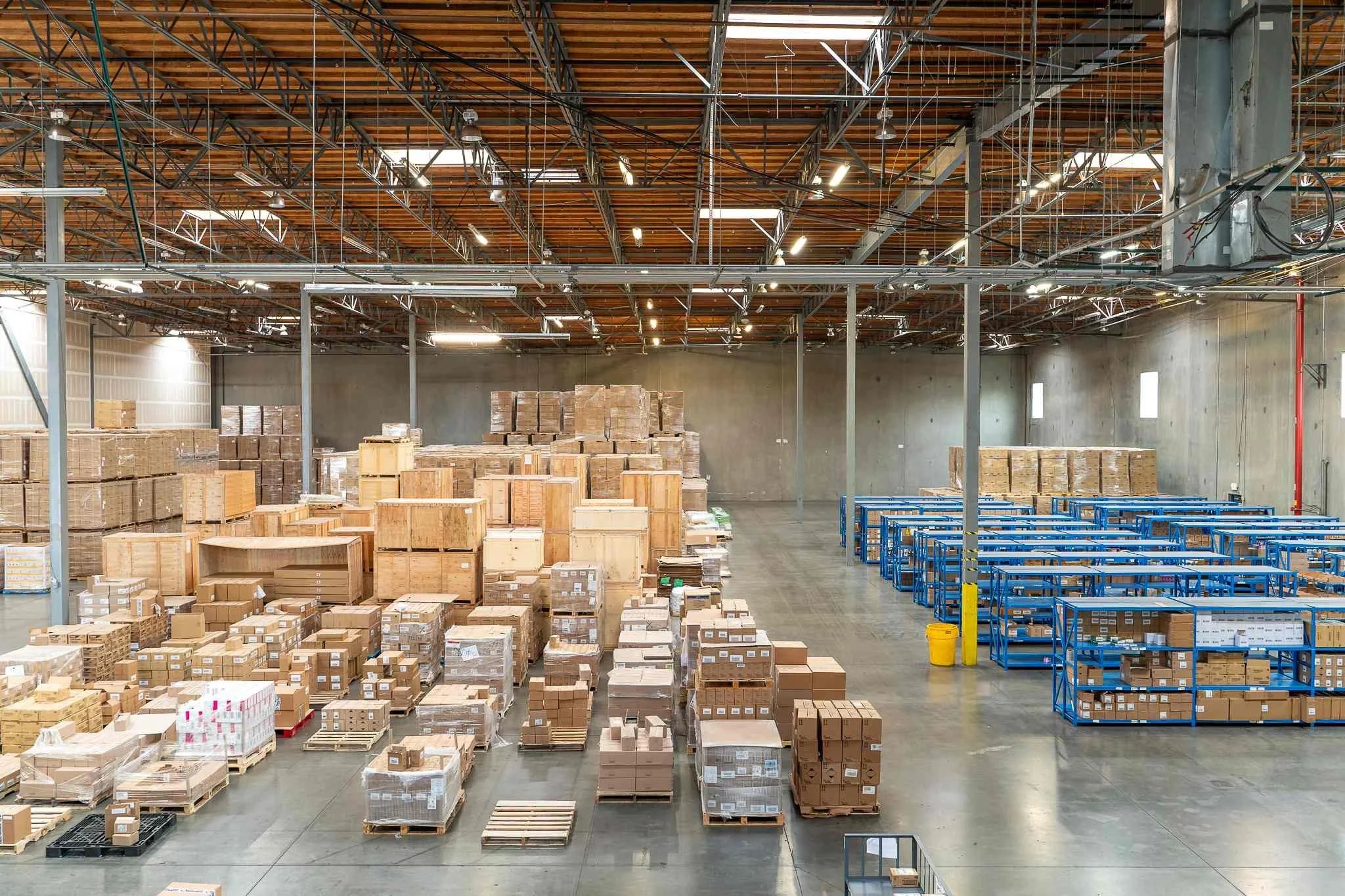 An interior view of a warehouse with stacked cardboard boxes on pallets and blue rolling carts, metal shelving, a wooden ceiling, and concrete walls.