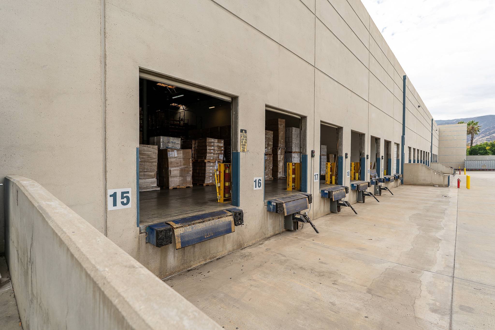 Warehouse loading dock with multiple open bays filled with stacked boxes and pallets, some wrapped in plastic, and numbered signs above each bay, with a mountain landscape and cloudy sky in the background.