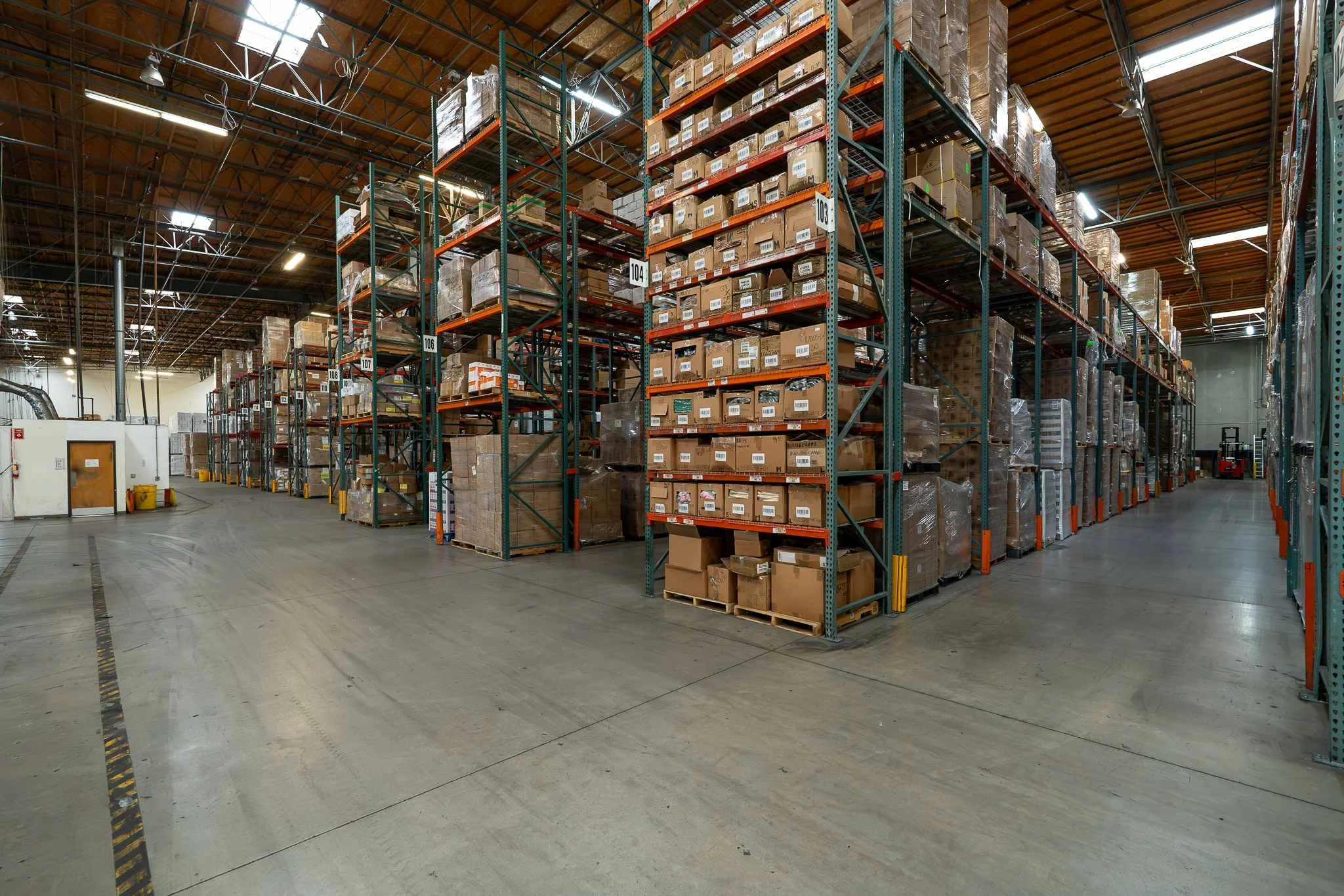 Interior of a warehouse with tall shelves stocked with boxes and pallets, concrete floor, and a wooden ceiling with metal beams and lighting.