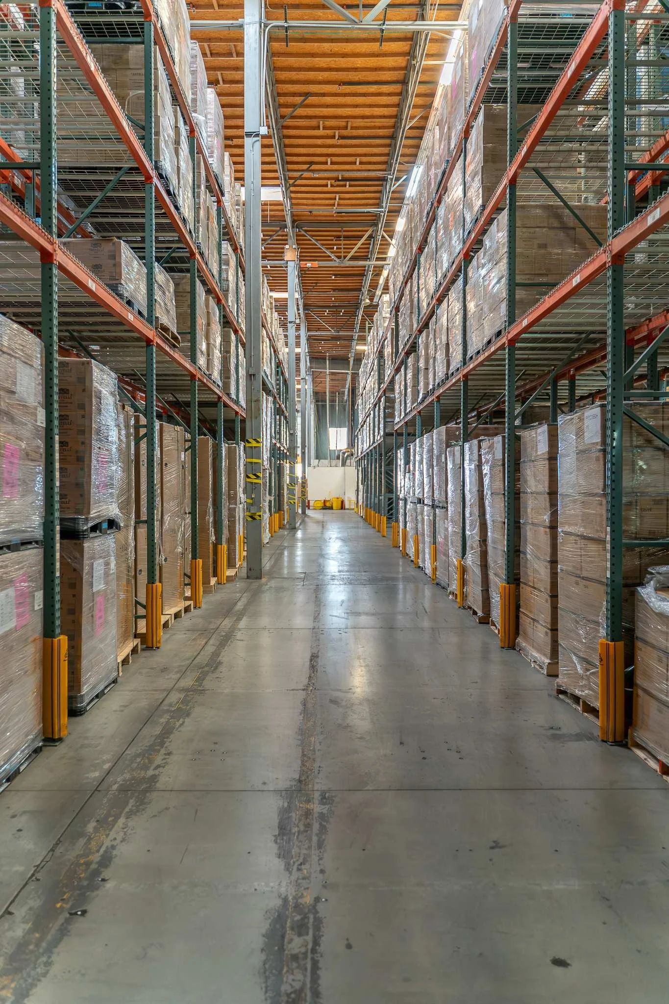 A large warehouse aisle with tall shelving units on both sides, filled with stacked boxes wrapped in plastic, and a concrete floor with visible tire marks.