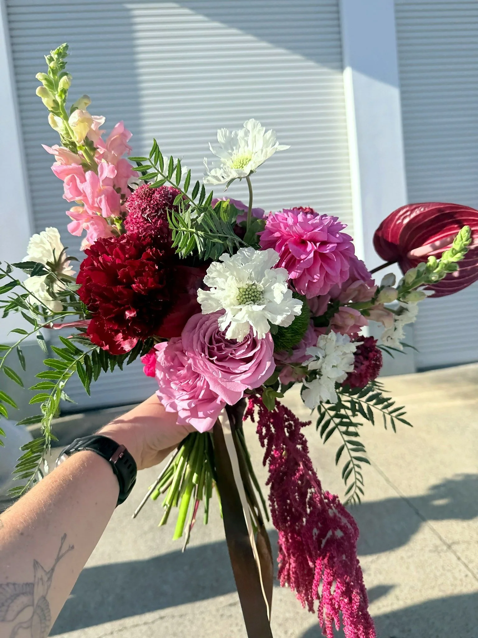A person holding a bouquet of pink, red, white, and purple flowers with green leaves outside in front of a building with slatted siding.