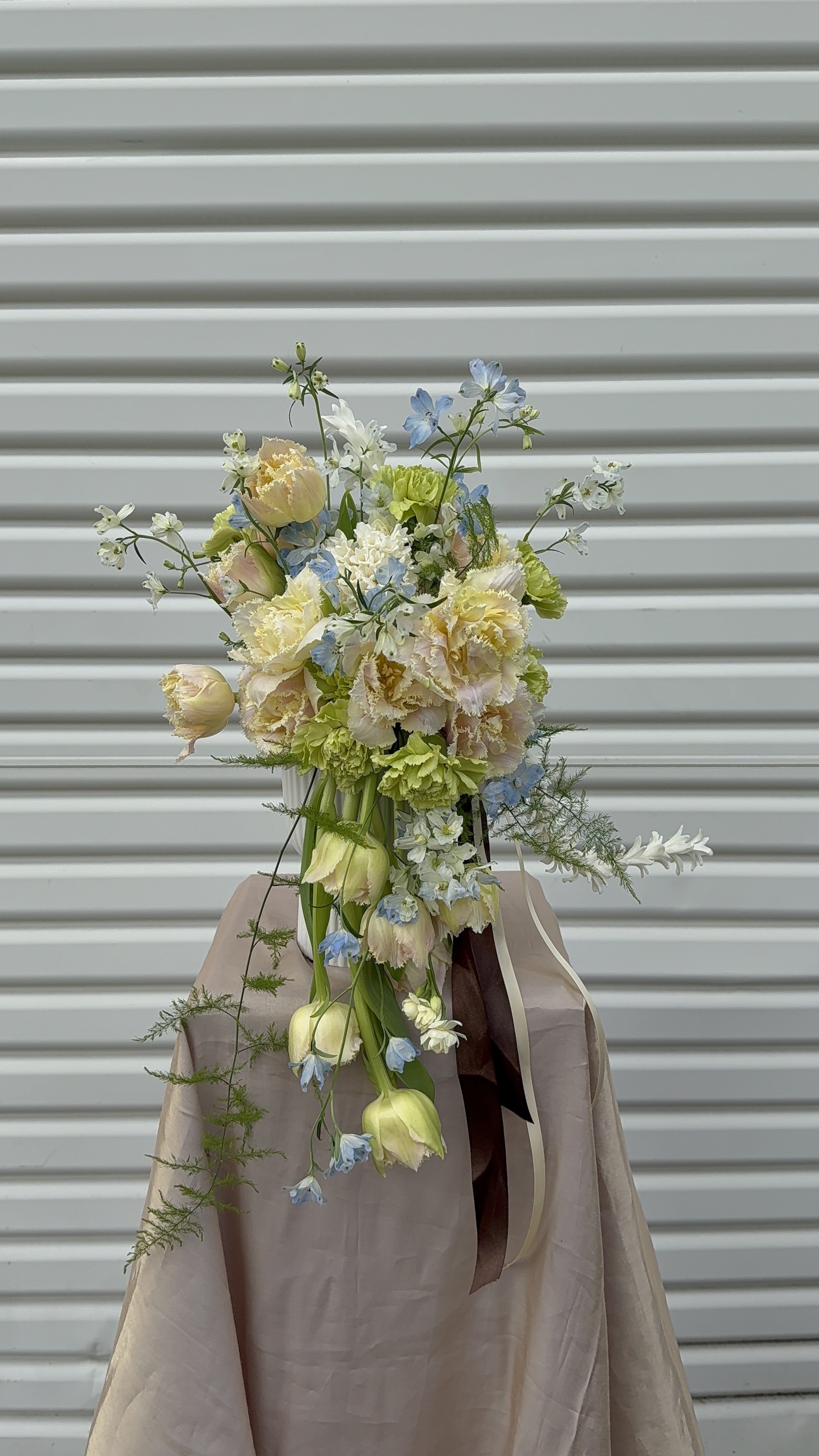 A floral bouquet with white, cream, and light blue flowers arranged on a beige tablecloth in front of a white, horizontal slatted background.