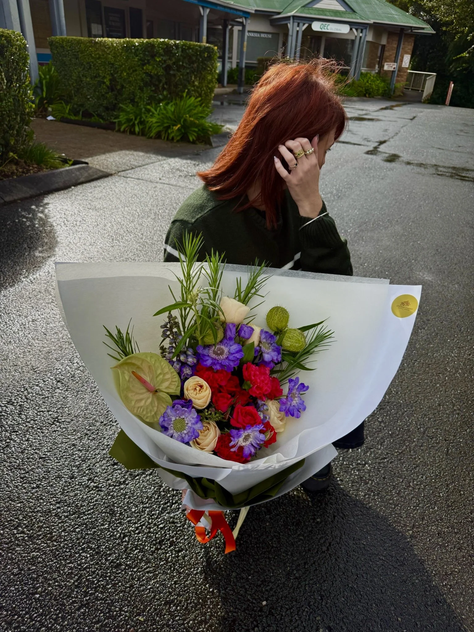 A woman with red hair holds a large bouquet of colorful flowers, including purple, red, orange, and cream-colored blooms, wrapped in white paper with a red ribbon. She is standing on a wet asphalt surface outside a building with a green roof and surrounding greenery.