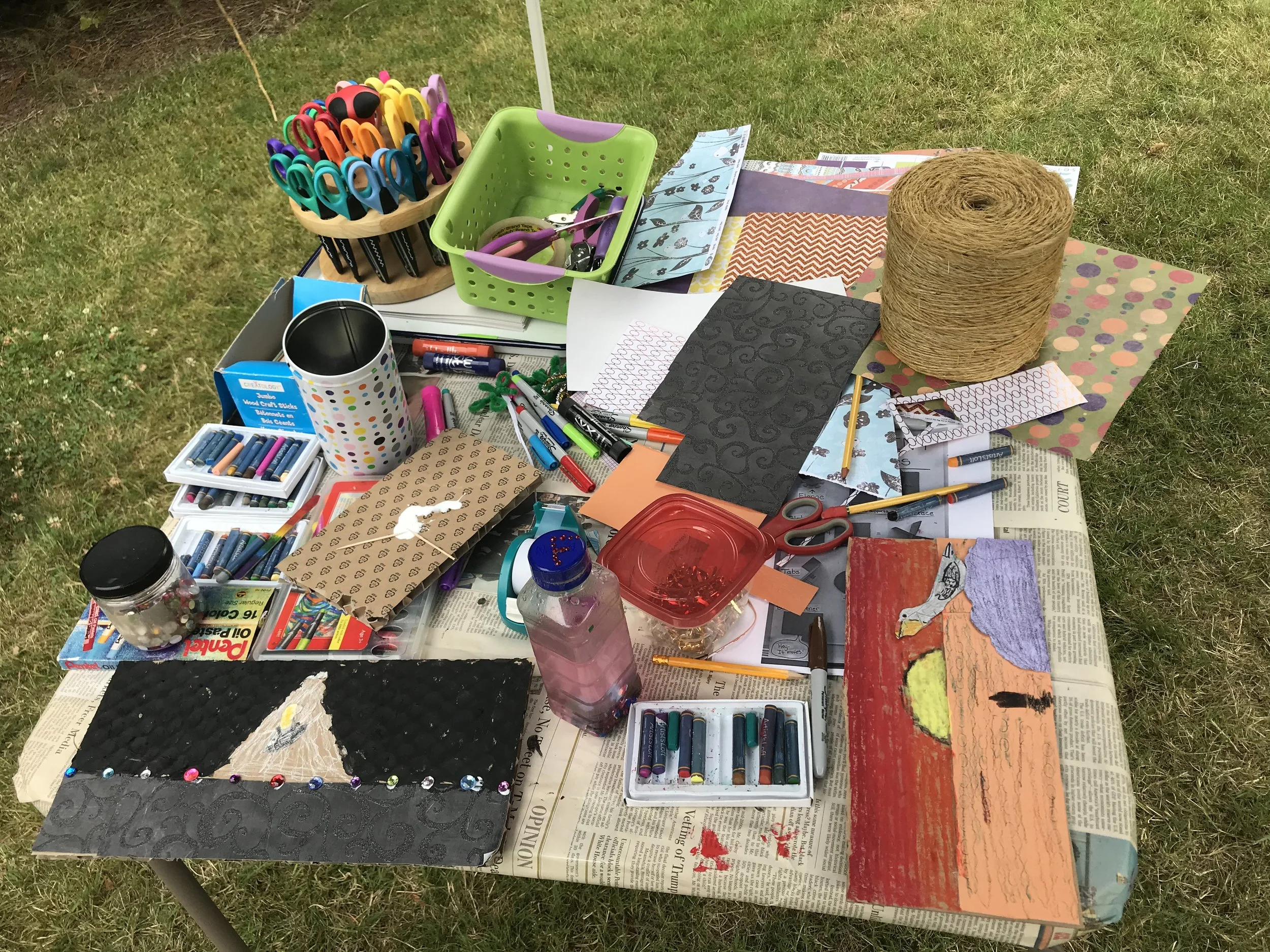 A table of art supplies (markers, paint, paper, yarn) set up outdoors for a class.