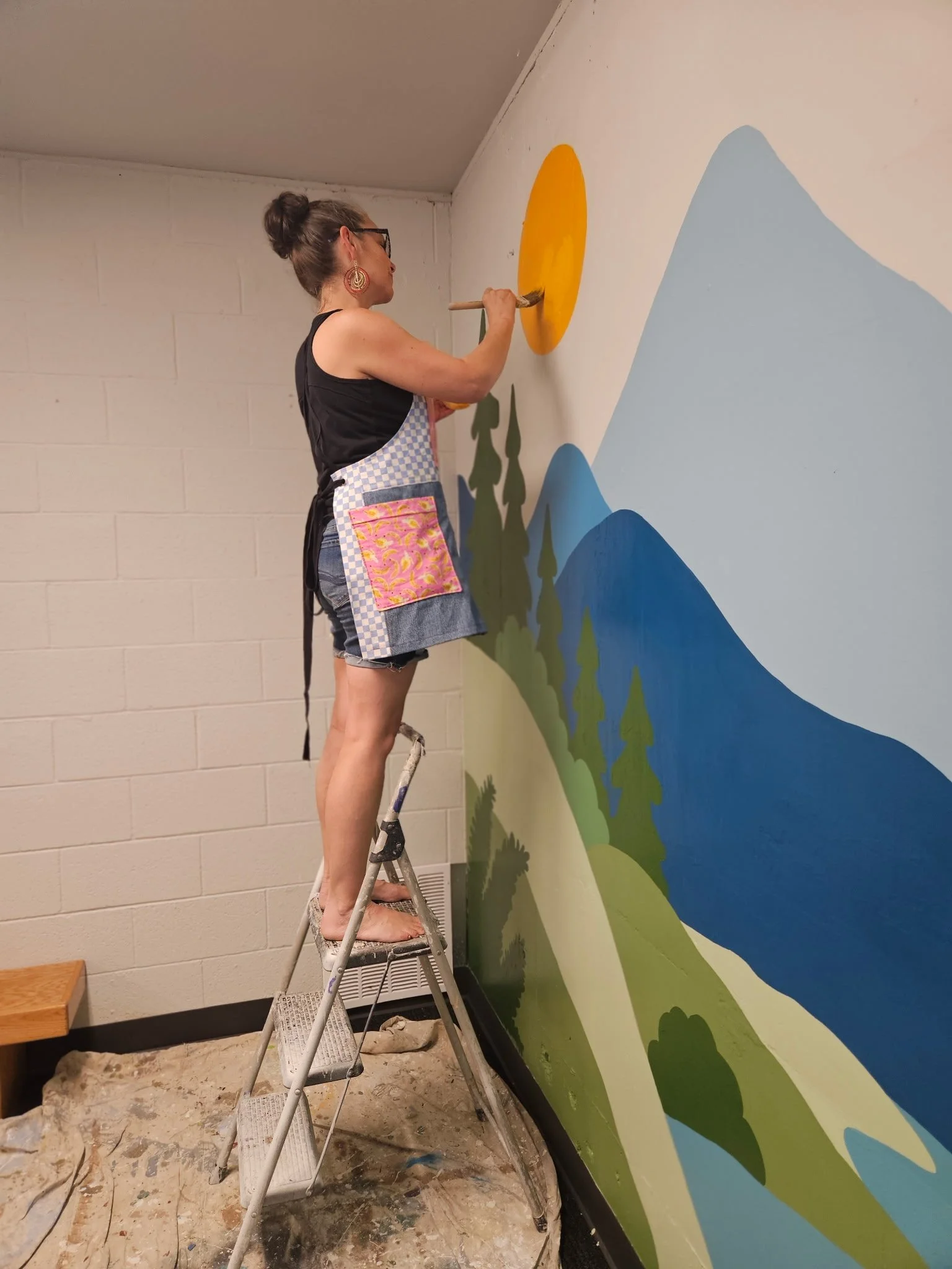 Artist Darcy Ballew on a step ladder, brush to the painted sun of a hand-painted mountain and forest landscape mural in Kirkland.