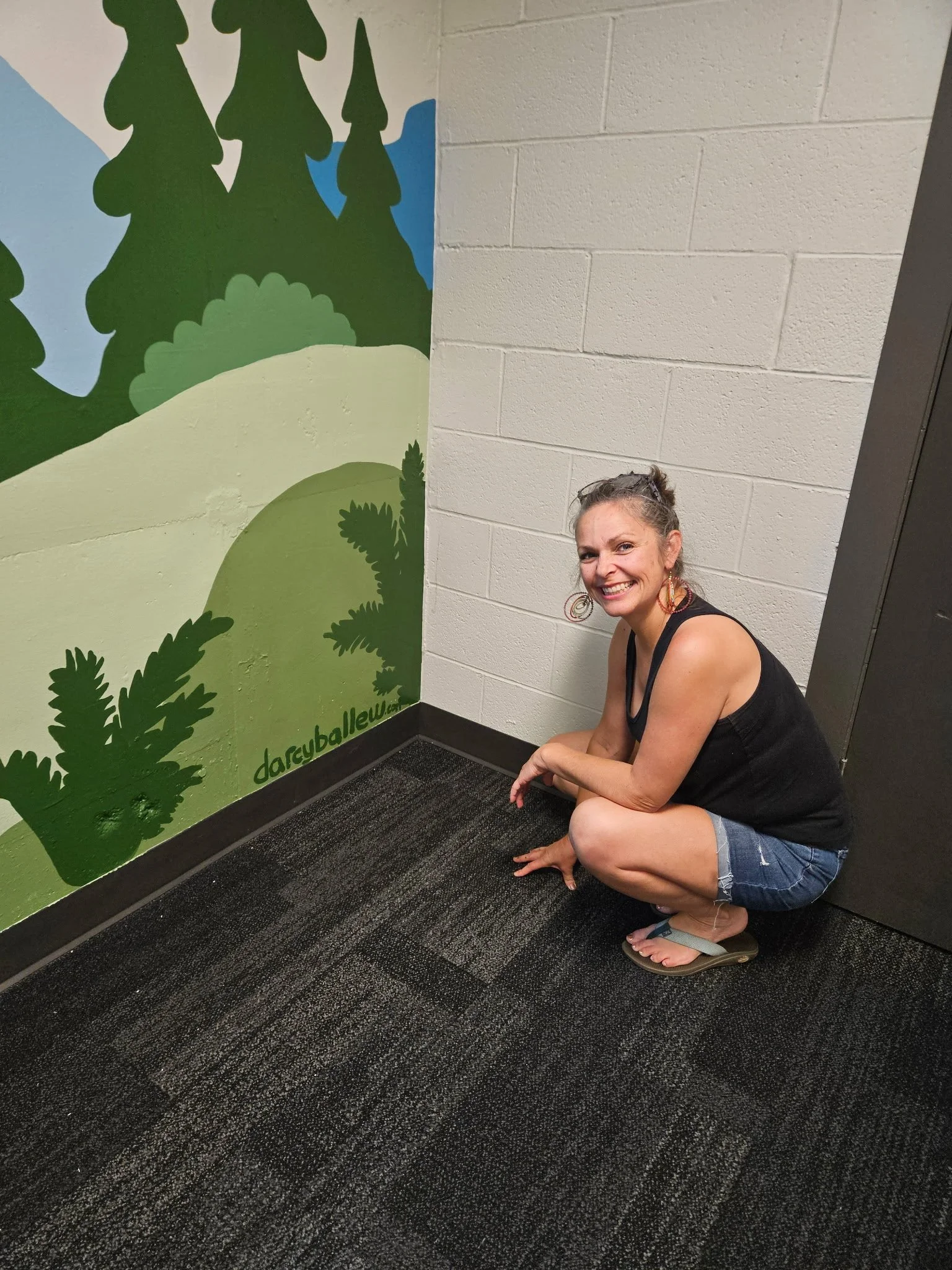 Darcy Ballew crouched beside her finished hand-painted forest and hills mural in Kirkland, pointing to her painted darcyballew.com signature.