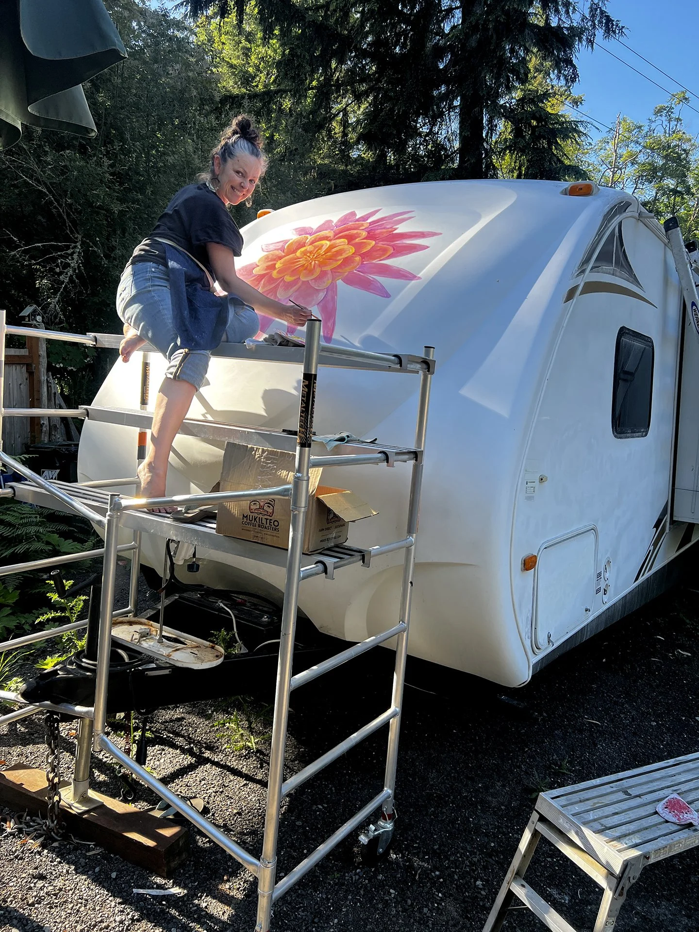 Muralist Darcy Ballew on scaffolding painting a large pink-and-orange dahlia on the front of a white RV at a yoga retreat.