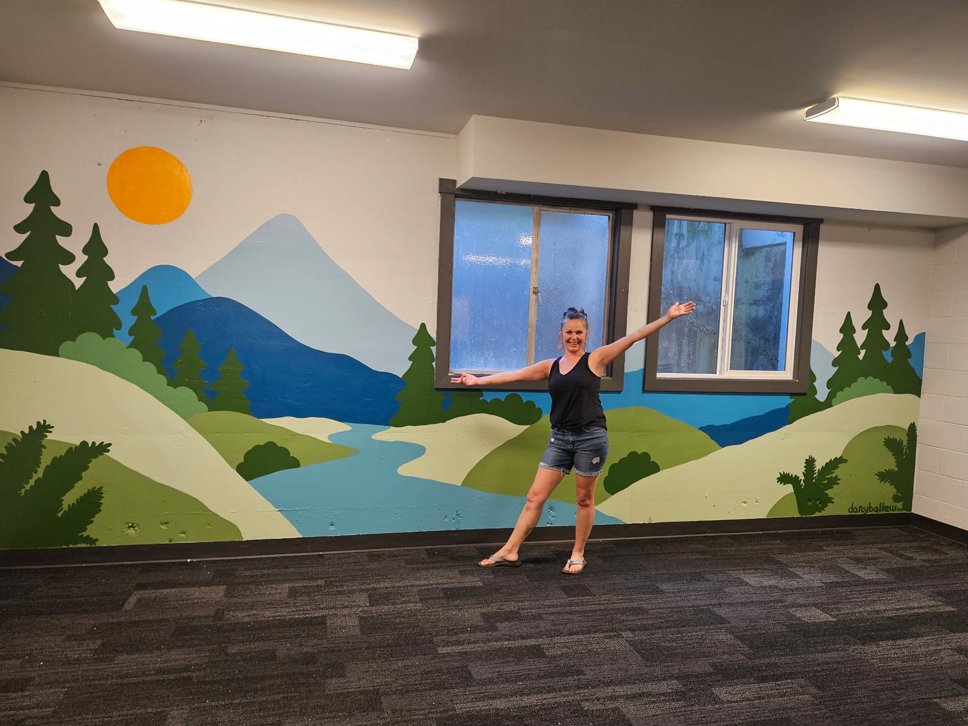 Hand-painted mountain and forest landscape mural by Darcy Ballew in a Kirkland community room, artist standing in front with arms out.