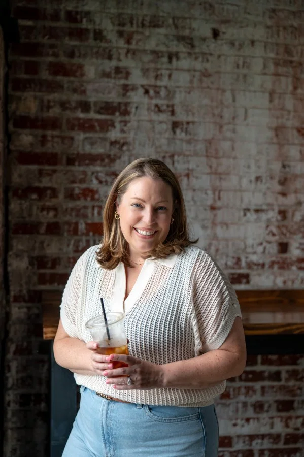 A smiling woman holding a glass of iced tea with a straw in front of a brick wall.