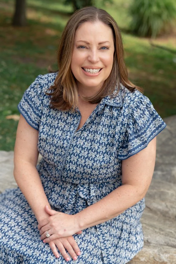 A woman with shoulder-length brown hair and blue eyes smiling outdoors, wearing a blue and white patterned dress with short sleeves, sitting on a rock with a grassy area and trees in the background.