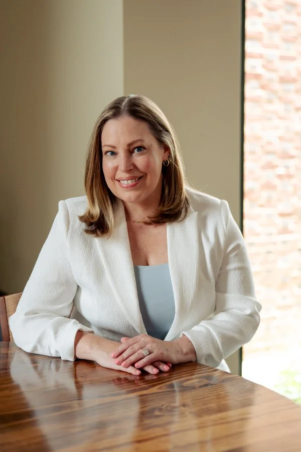 A smiling woman with shoulder-length hair wearing a white blazer and light-colored top, sitting at a wooden table indoors with a window in the background.