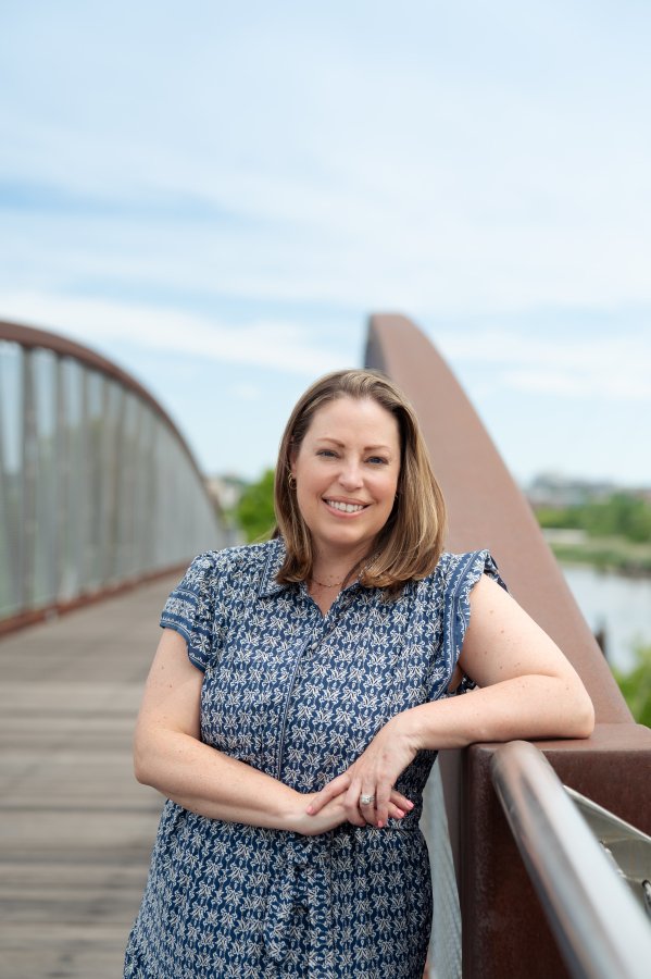 Woman smiling on a bridge outdoors in daytime, wearing a patterned blue dress.