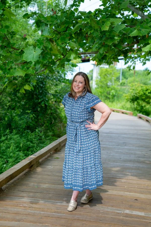 A woman in a blue patterned dress standing on a wooden walkway surrounded by green trees and bushes.