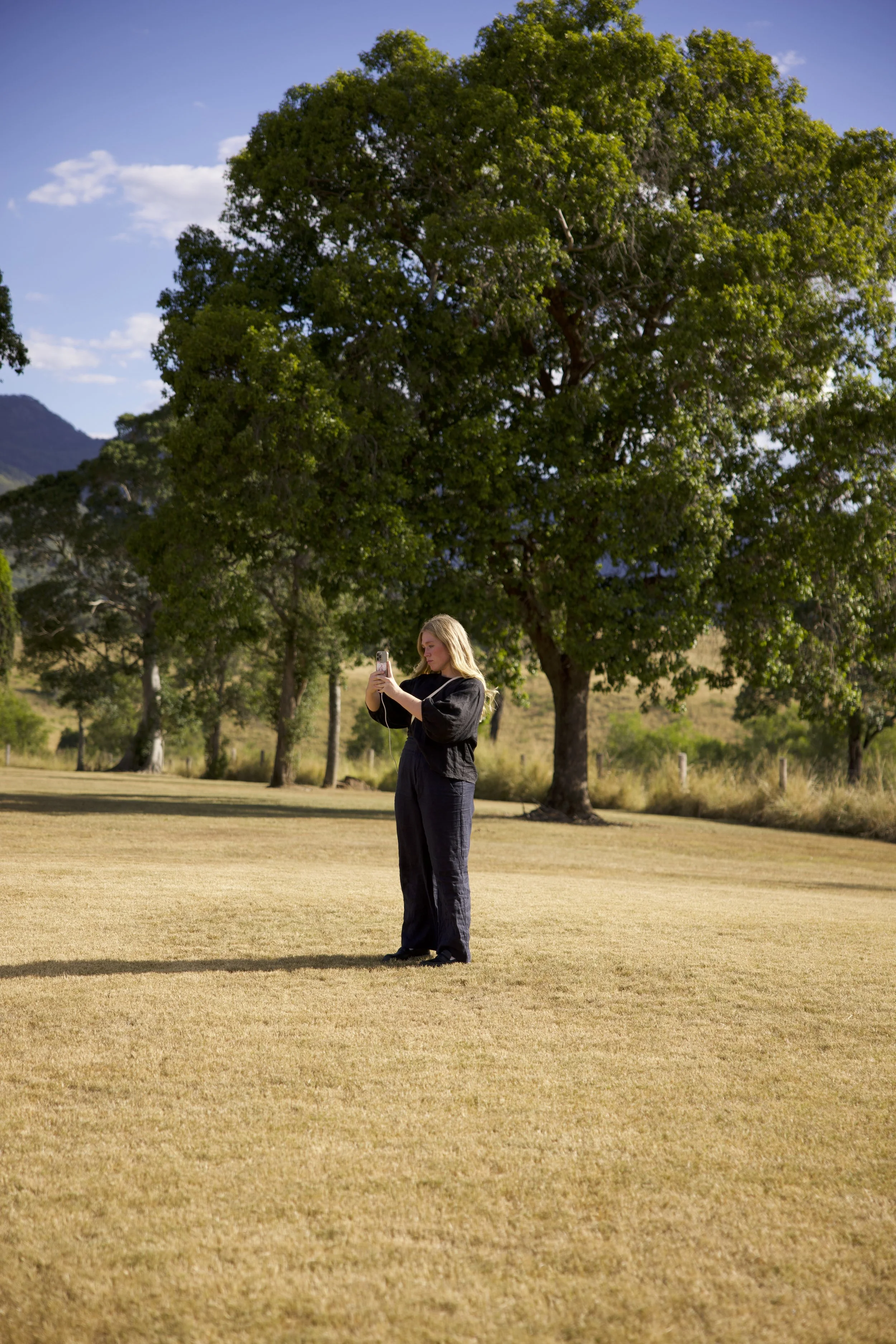A woman standing on a dry grassy field, taking a photo with her phone. Large trees and mountains are in the background, with a mostly clear sky.