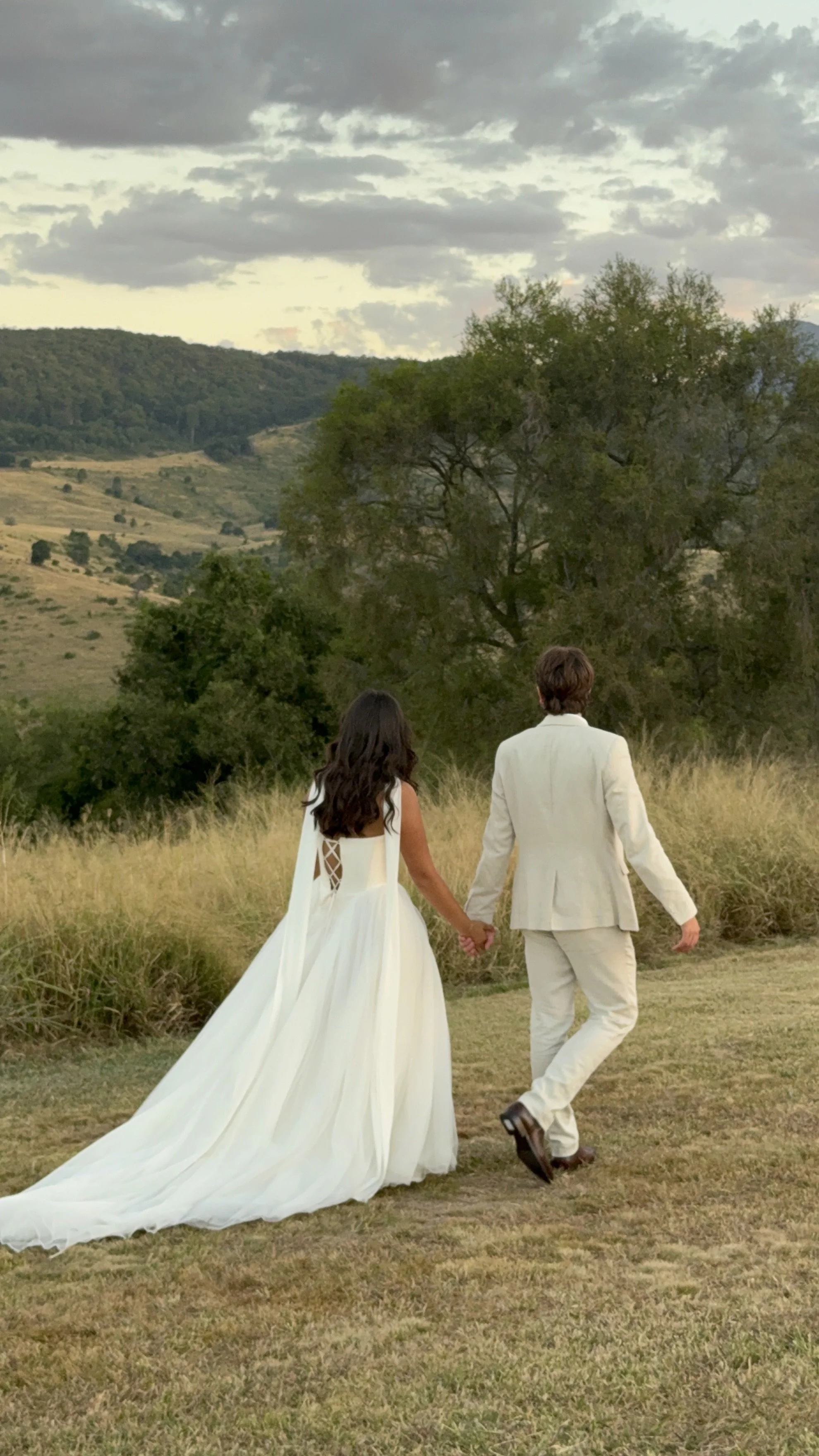 A bride and groom walking hand-in-hand in a grassy field with mountains and trees in the background, under cloudy sky.