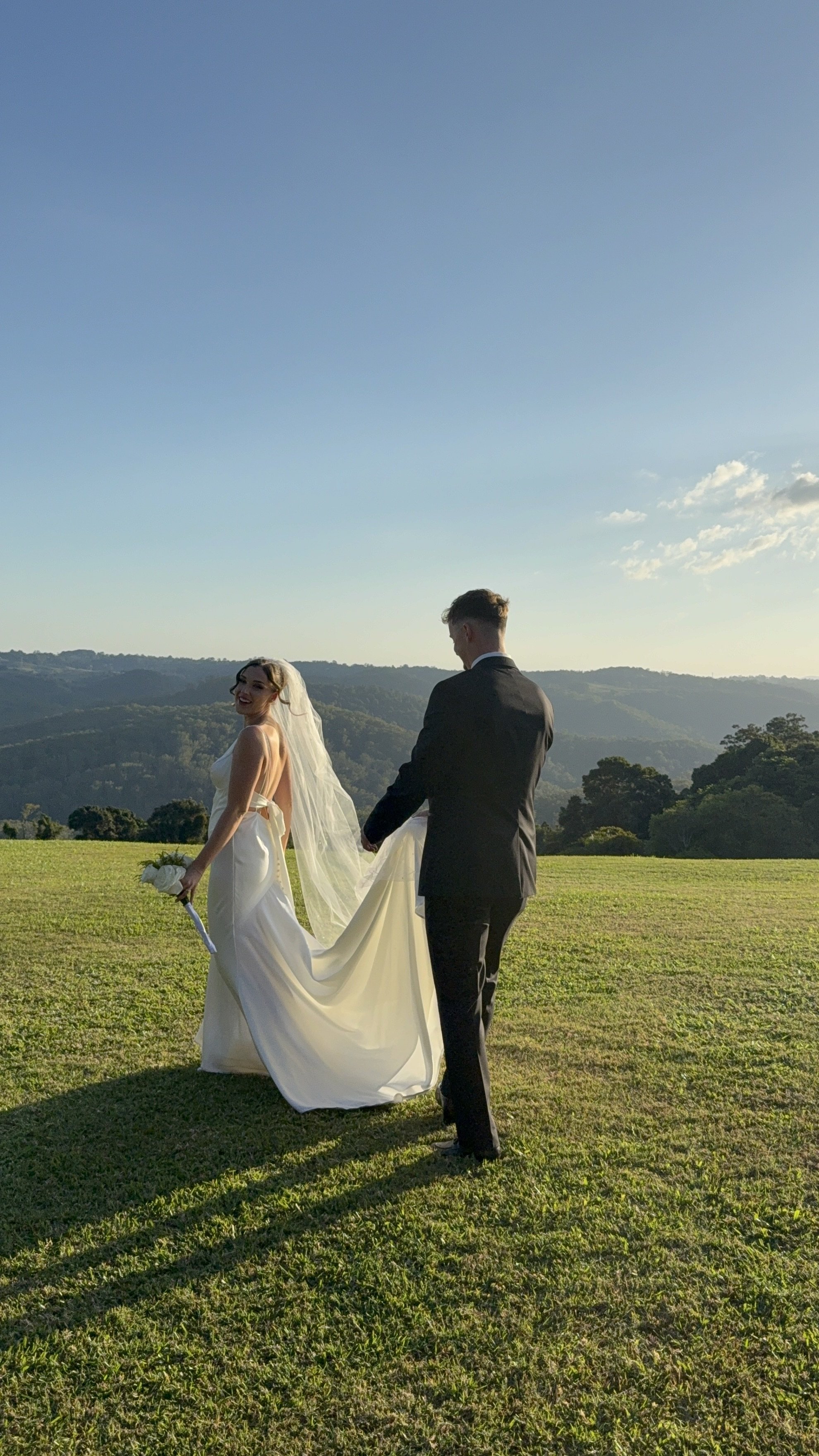 A bride and groom holding hands outdoors on a sunny day with green grass and hills in the background, the bride smiling and holding a bouquet.