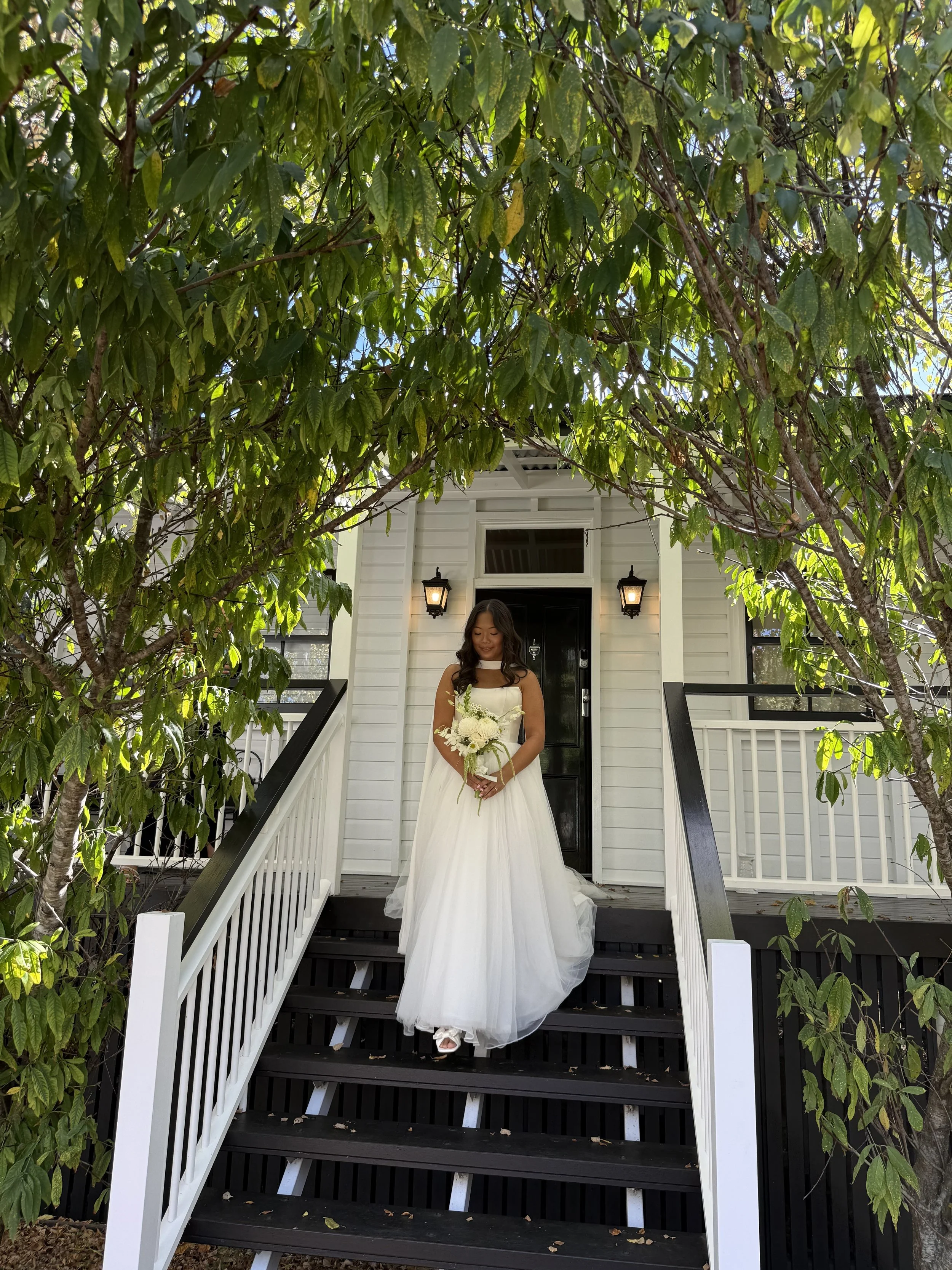 A bride standing on the steps of a white house with black door and lanterns, surrounded by green trees, holding a white bouquet and smiling.
