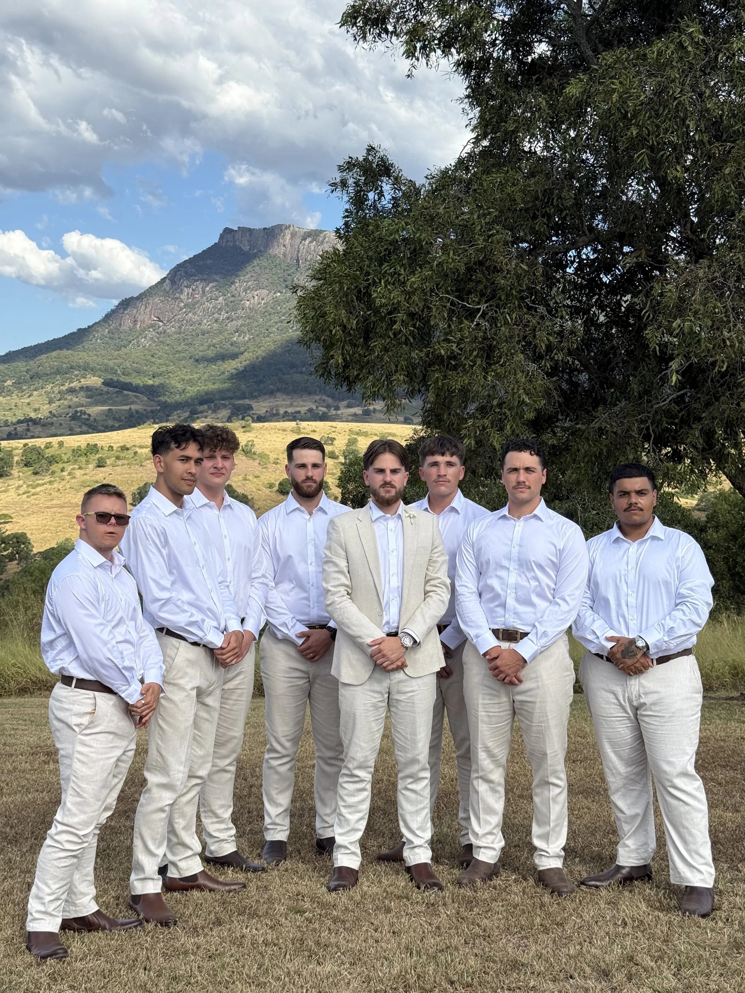 A group of nine men dressed in white shirts and light-colored pants, standing outdoors on grass with a mountainous landscape and partly cloudy sky in the background.