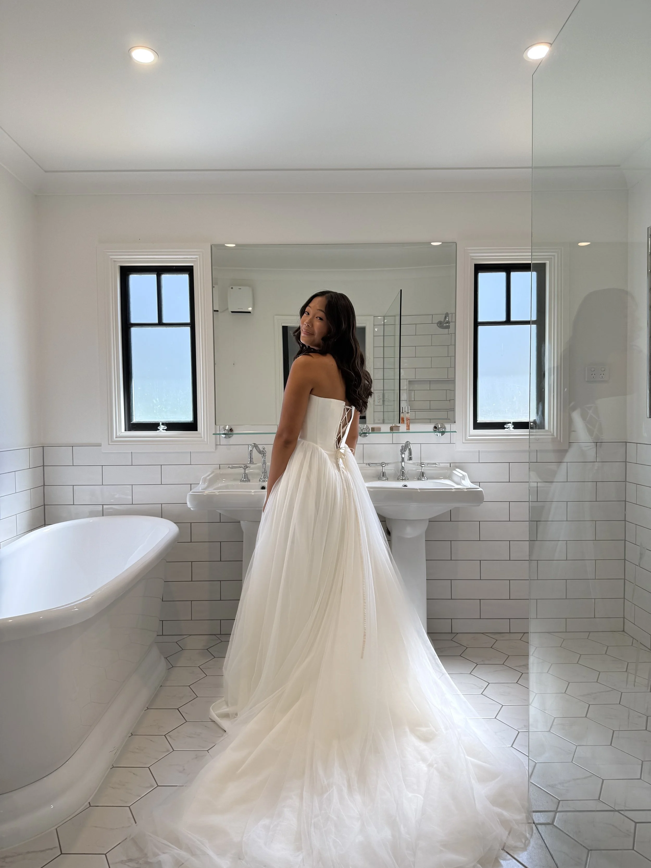 A woman in a white strapless wedding gown standing in a bright bathroom with white hexagonal tile on the floor and walls, and black-framed windows. She is facing away but turning her head to smile at the camera, with long, dark, wavy hair and minimal makeup.