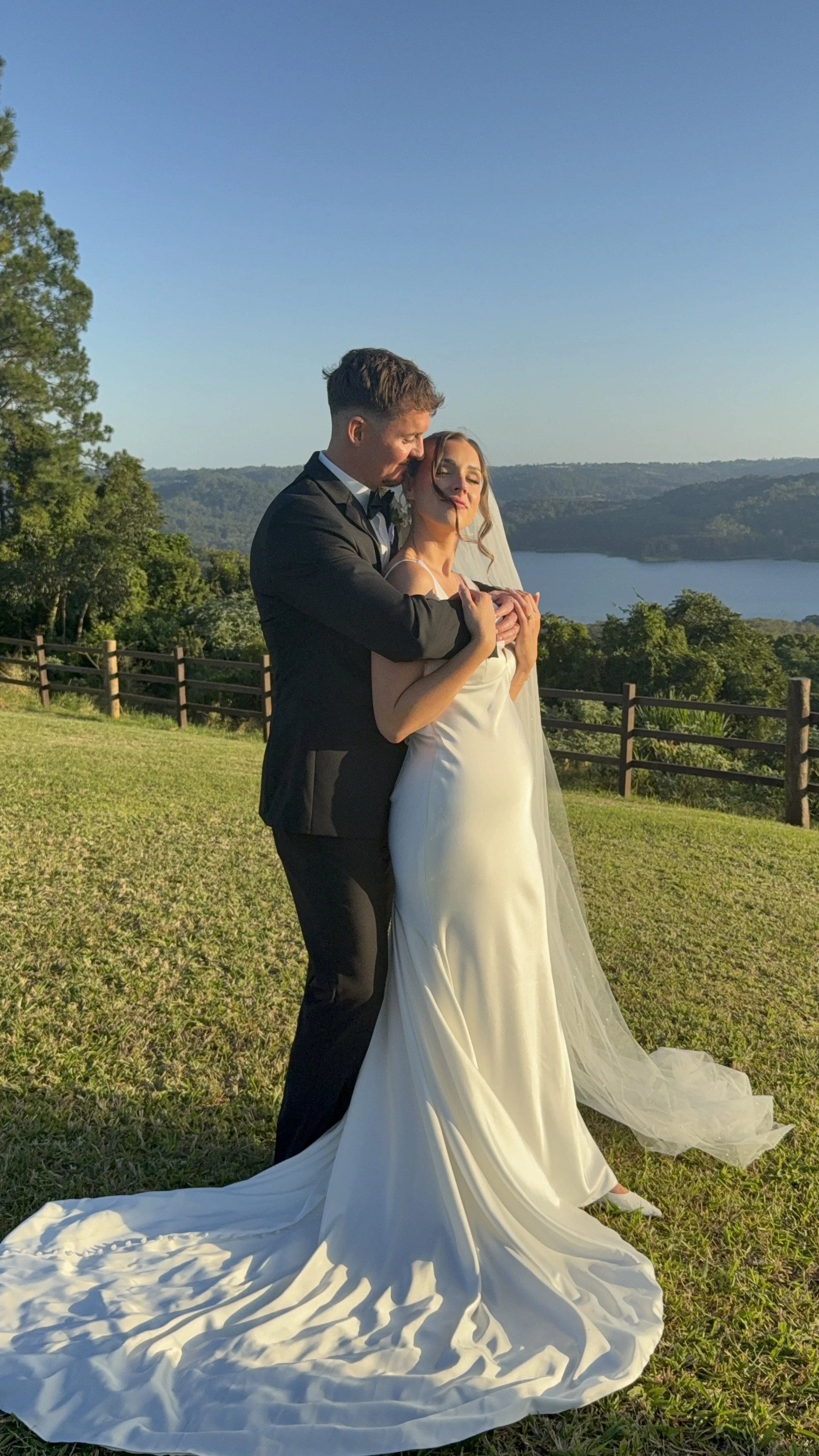A bride and groom embracing outdoors on a grassy hill with a lake and hills in the background.