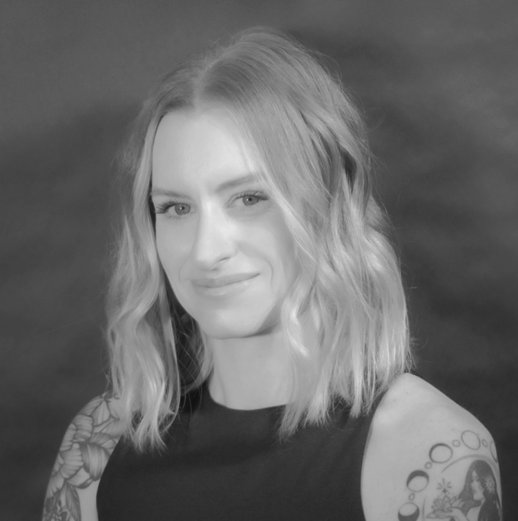 Black and white portrait of a woman with wavy shoulder-length hair, smiling slightly, wearing a sleeveless top, with a tattoo on her right arm.