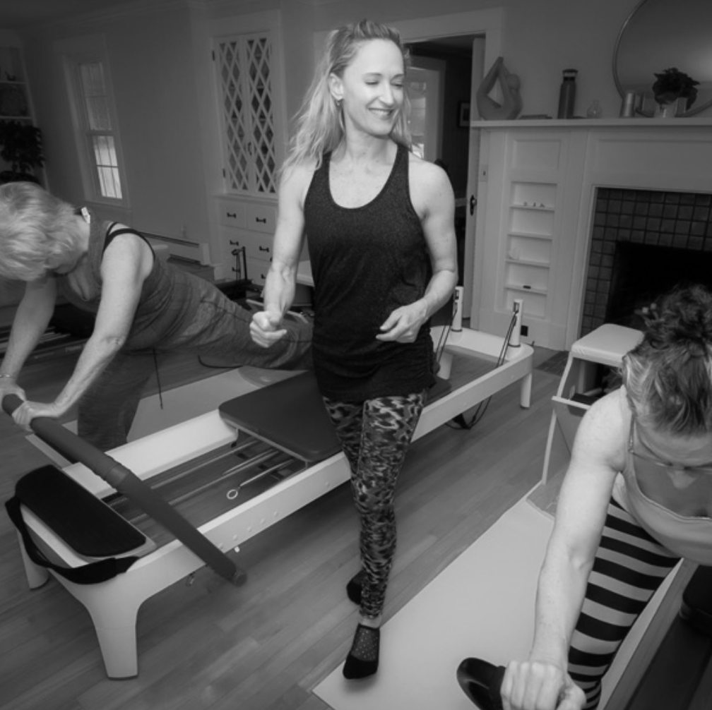 Three women exercising indoors, one on a Pilates reformer machine, one doing a plank, and one holding a barbell in a living room.