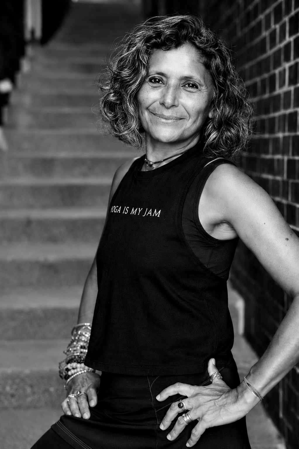 A smiling middle-aged woman with curly hair standing outdoors next to a brick wall and stairs, wearing a sleeveless shirt that reads 'YOGA IS MY JAM' and jewelry.