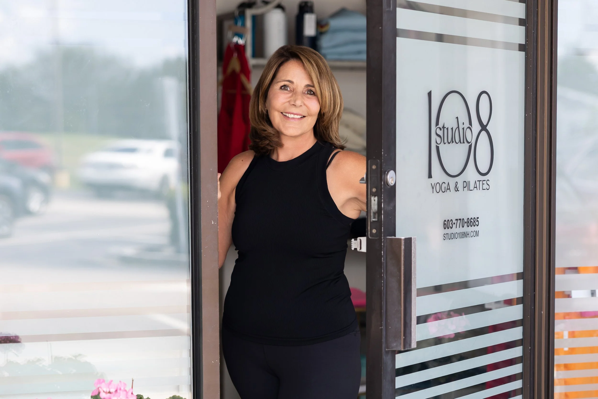 A smiling woman with shoulder-length brown hair standing in a doorway of a yoga studio, with the studio's name and contact information on the glass door.