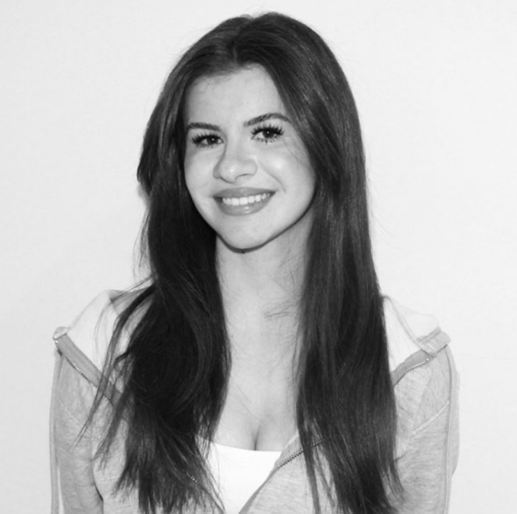 Black-and-white photo of a young woman with long dark hair, smiling at the camera, wearing a light-colored jacket over a white top.