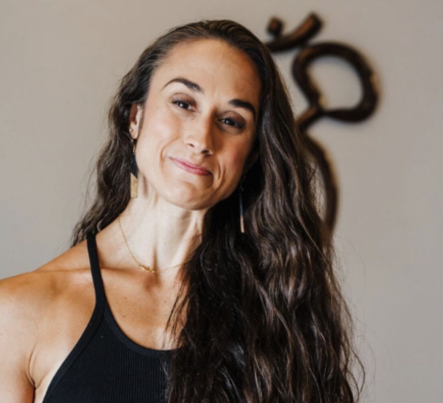 A woman with long wavy brown hair smiling at the camera, wearing a black tank top and gold jewelry, with artwork in the background.
