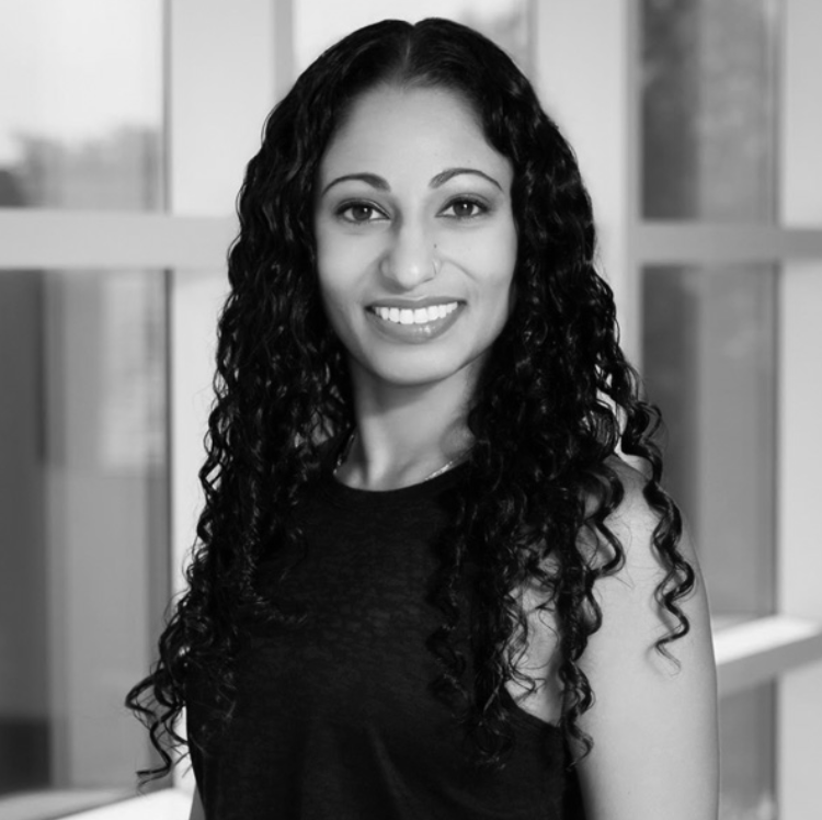 Black and white photograph of a smiling woman with long, curly hair, wearing a sleeveless top, standing in front of a window.