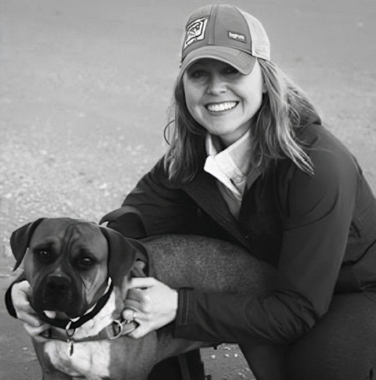 A woman wearing a baseball cap, jacket, and smiling, crouches next to a large dog with a collar, on an outdoor surface.