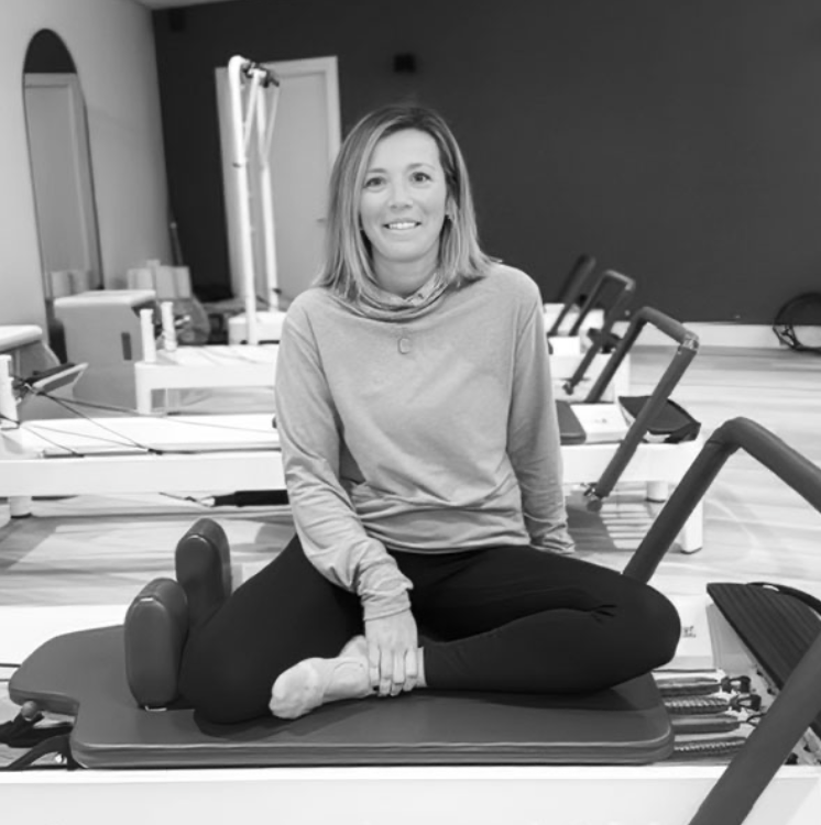A woman in activewear sitting cross-legged on a Pilates reformer machine in a fitness studio, smiling at the camera.