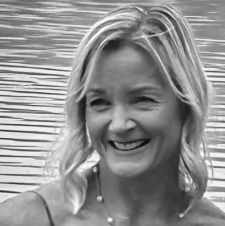 A smiling woman with wavy hair standing near a body of water with horizontal ripples.