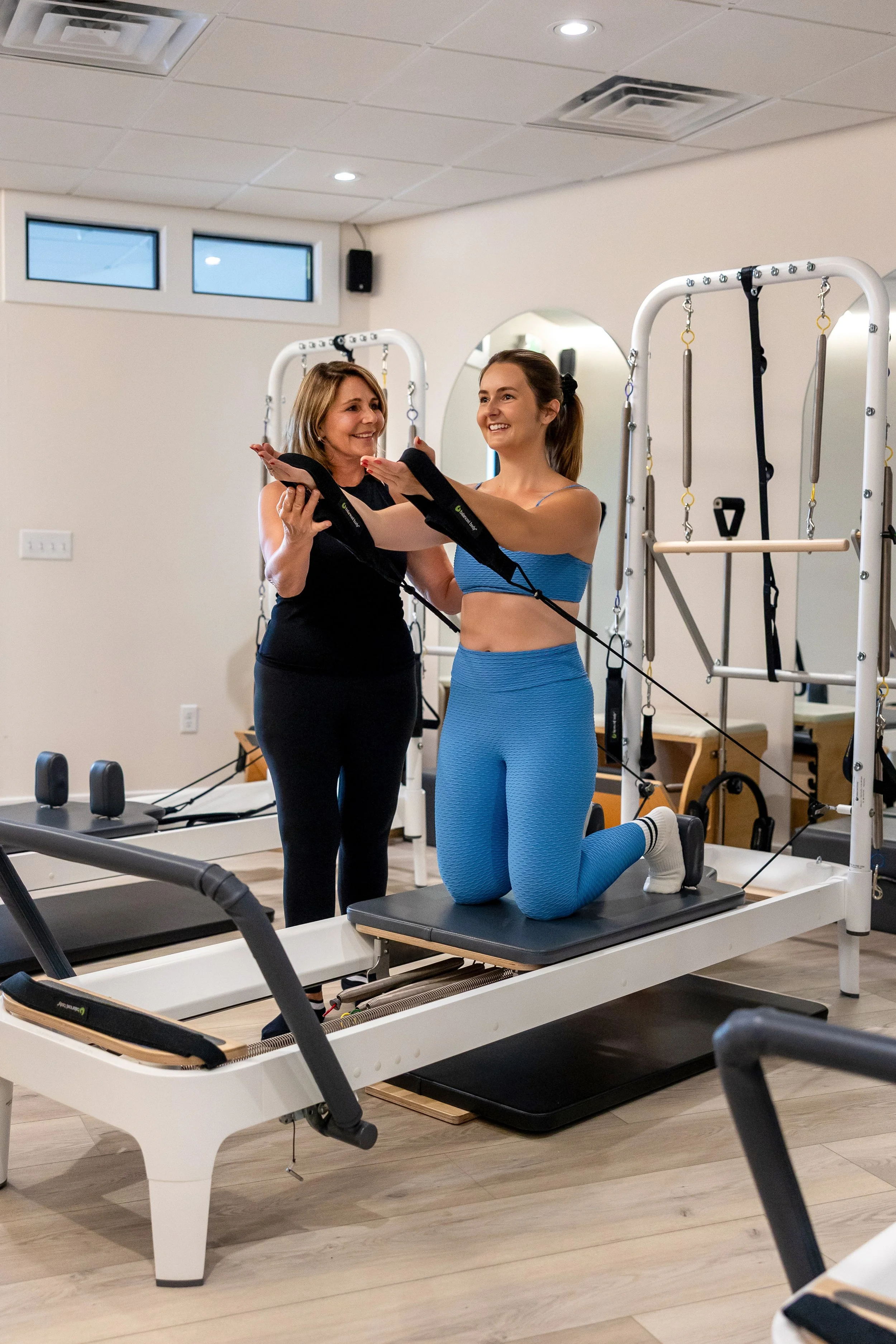 A woman receiving a Pilates or physical therapy exercise from an instructor in a bright, modern studio with Pilates reformer equipment and large windows.
