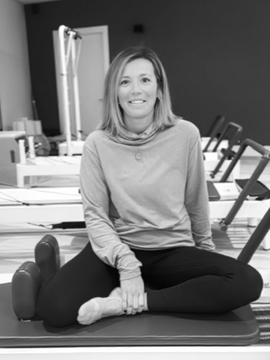 A woman sitting cross-legged on a yoga mat in a Pilates or yoga studio, smiling at the camera, with exercise equipment and mirrors in the background.