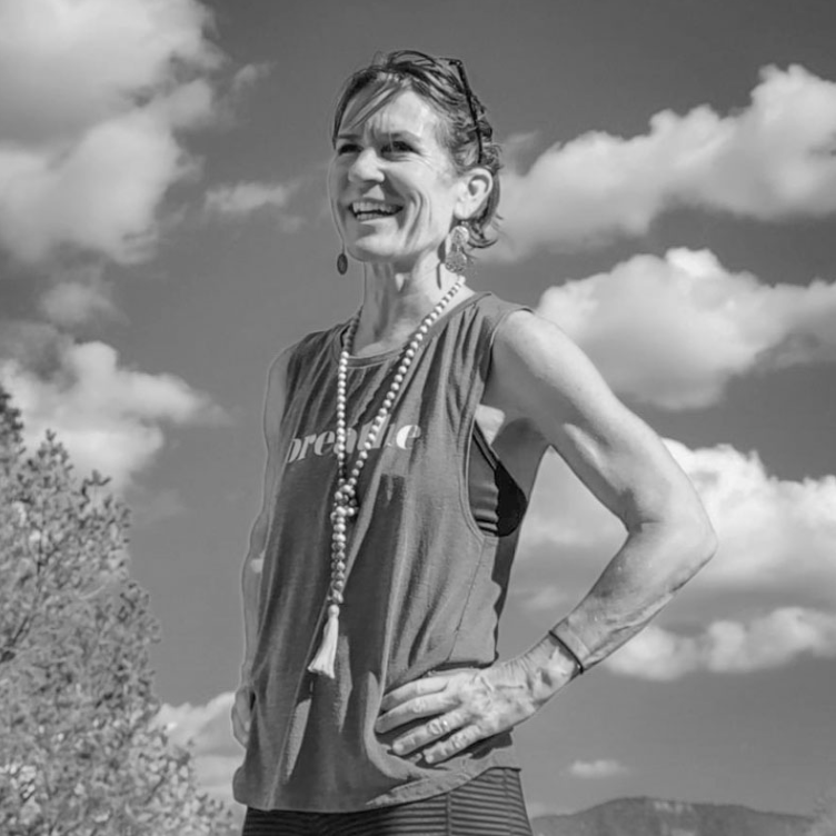 A smiling woman standing outdoors with her hand on her hip, wearing athletic clothing and a beaded necklace, under a partly cloudy sky.