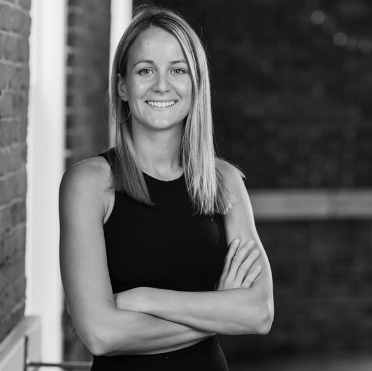 Black and white portrait of a woman with shoulder-length hair, wearing a sleeveless top, smiling, standing outdoors with arms crossed, brick wall in the background.